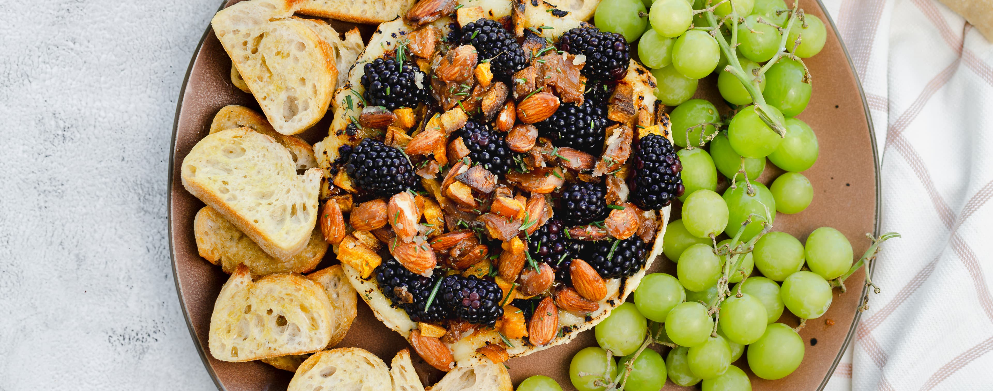 A plate filled with a Brie brûlée with butternut squash and toffee almonds appetizer, accompanied by grapes and toasted bread.